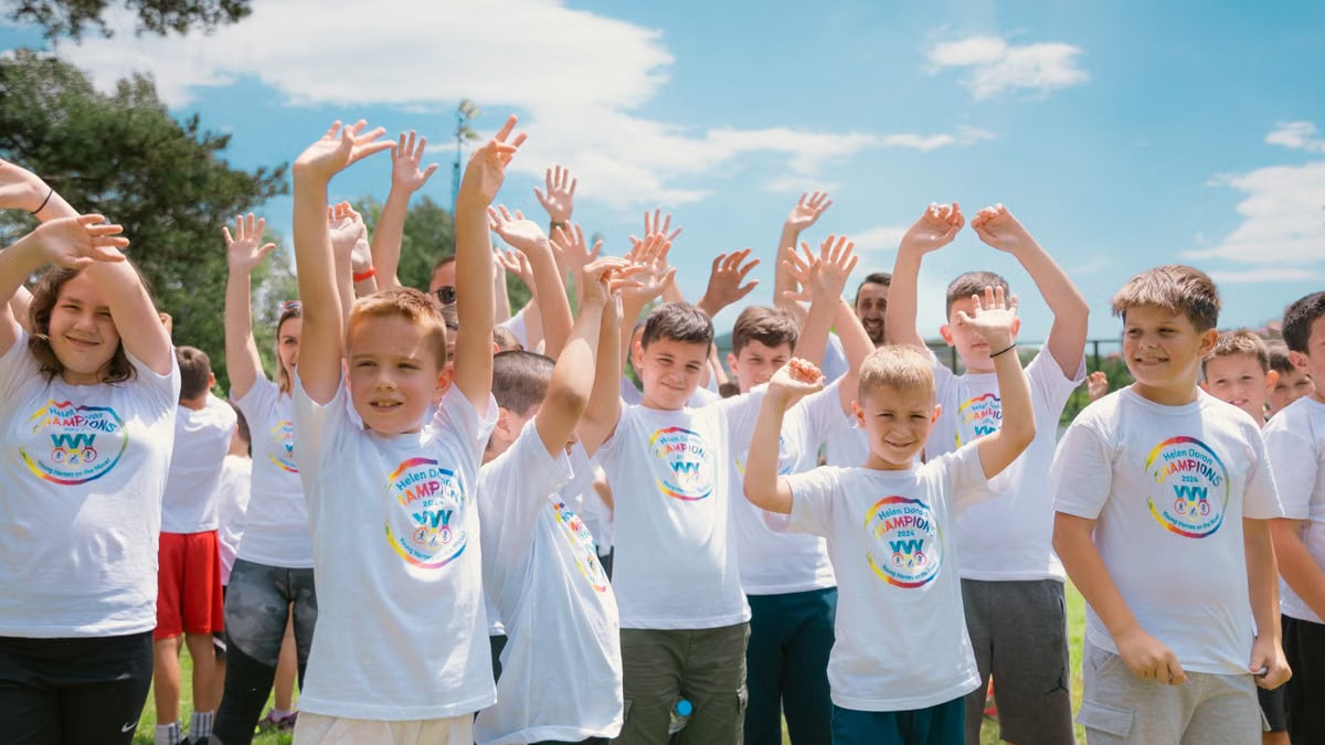 A group of cheerful children in white T-shirts raising hands in a park during summer.